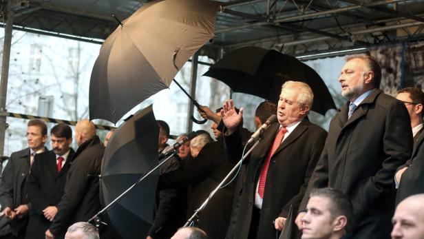 Miloš Zeman spricht auf einer Bühne, geschützt durch Regenschirme.