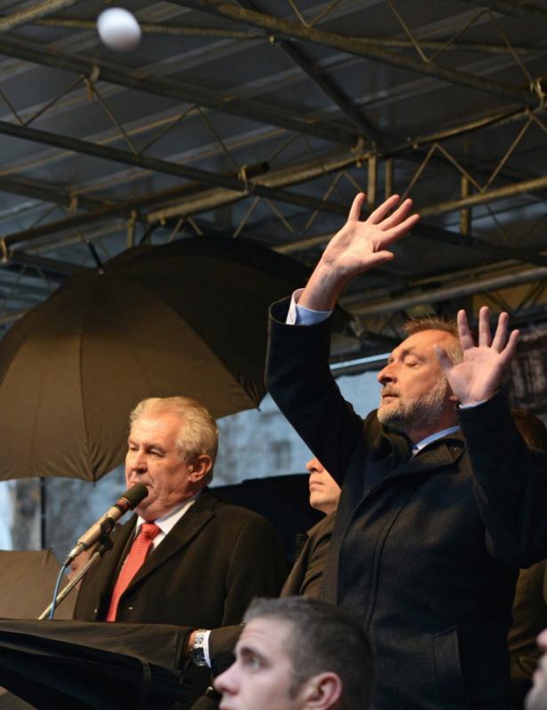 Ein Mann auf einer Bühne wehrt ein fliegendes Ei ab, während ein anderer ins Mikrofon spricht.