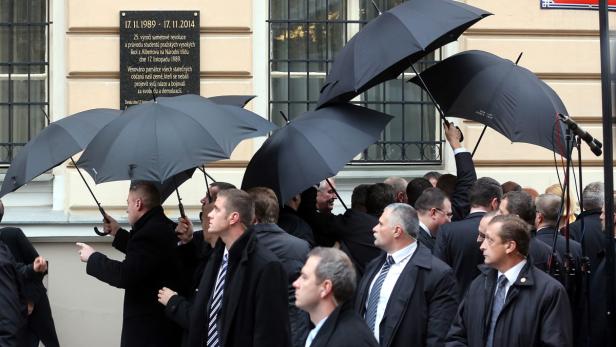 Eine Menschenmenge mit schwarzen Regenschirmen steht vor einer Gedenktafel.