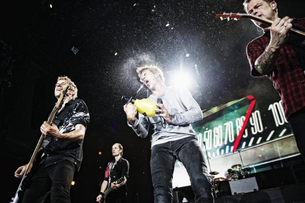 Eine Band tritt auf der Bühne auf, wobei ein Mitglied sich mit einer Sprühflasche Wasser ins Gesicht sprüht.