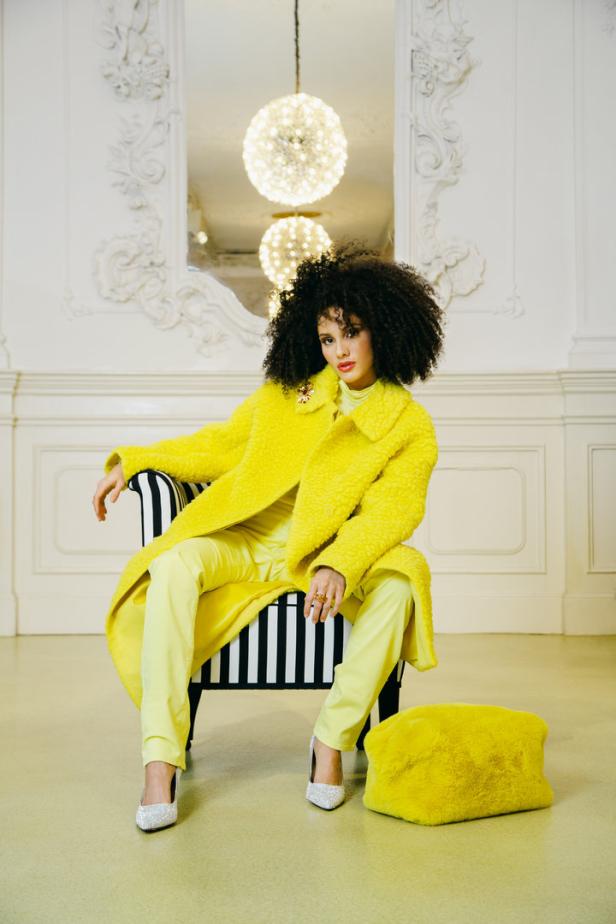 Eine Frau mit Afro sitzt in einem schwarz-weiß gestreiften Sessel und trägt einen gelben Mantel und glitzernde Schuhe.