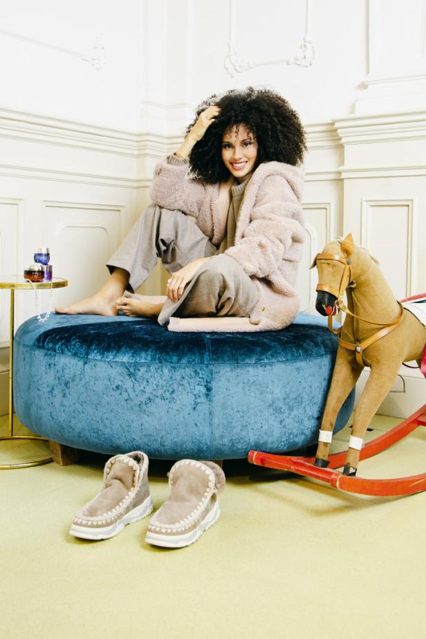 Eine Frau mit Afro sitzt barfuß auf einem blauen Samtpouf neben einem Schaukelpferd.