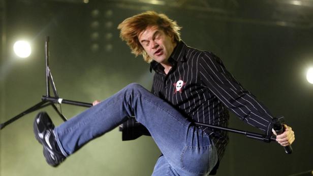 Ein Mann mit blonden Haaren springt mit einem Mikrofonständer auf der Bühne.