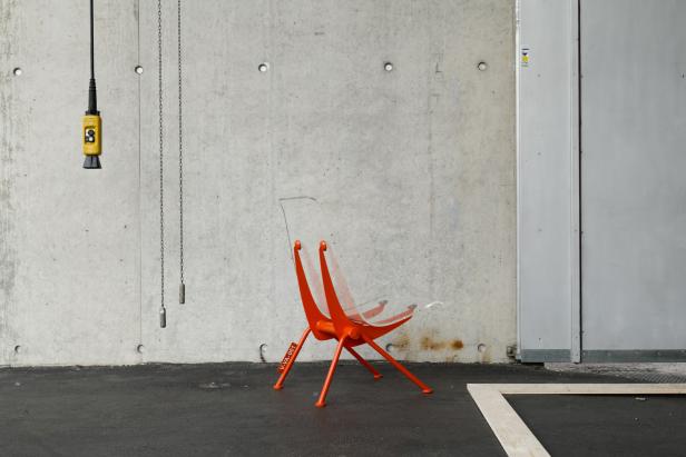 Ein orangefarbener Stuhl mit durchsichtiger Sitzfläche steht vor einer Betonwand.