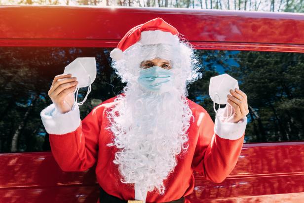 Ein Weihnachtsmann mit Mundschutz hält zwei weitere Masken in der Hand.