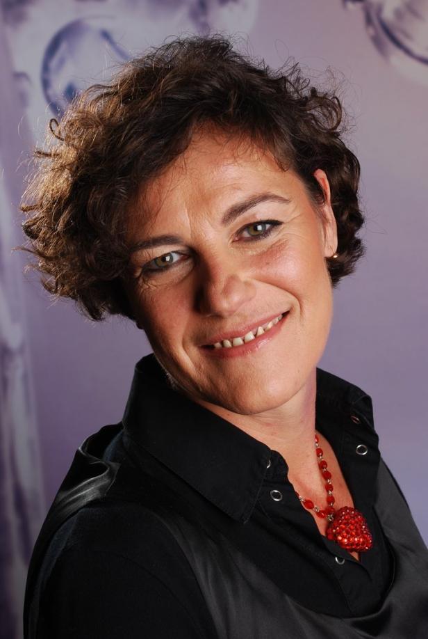 Eine lächelnde Frau mit kurzem, lockigem Haar und einer roten Halskette vor einem lila Hintergrund.