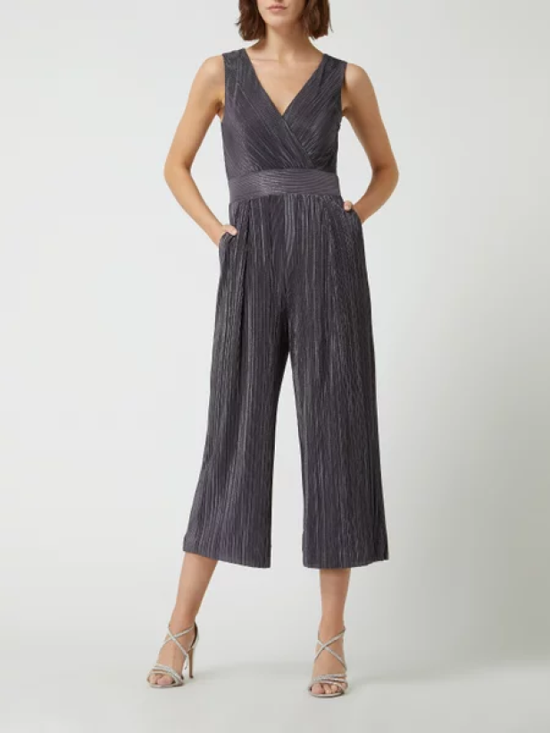 Eine Frau in einem grauen, plissierten Jumpsuit mit V-Ausschnitt und silbernen High Heels.