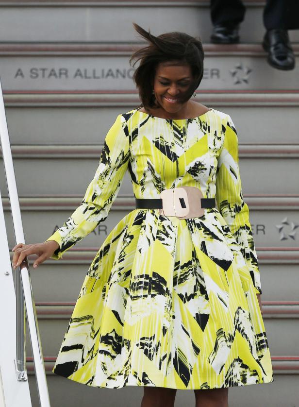 Michelle Obama geht eine Flugzeugtreppe in einem gelb-schwarzen Kleid hinunter.