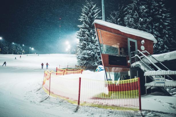 Nächtliche Aufnahme einer Skipiste mit Skifahrern und einem kleinen Gebäude im Vordergrund.