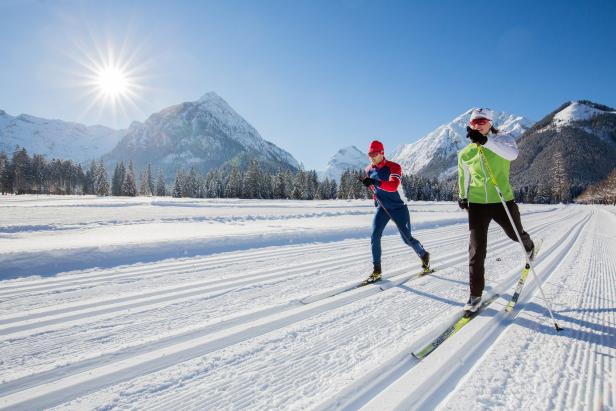 Zwei Langläufer auf einer Loipe vor einer verschneiten Berglandschaft.