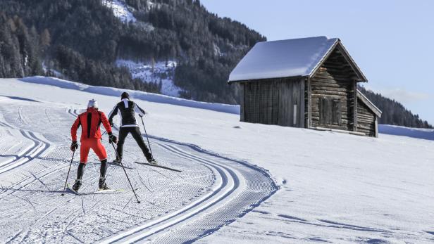 Zwei Langläufer auf einer Loipe vor einer schneebedeckten Hütte.