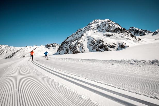 Zwei Langläufer auf einer Loipe vor einer verschneiten Berglandschaft.