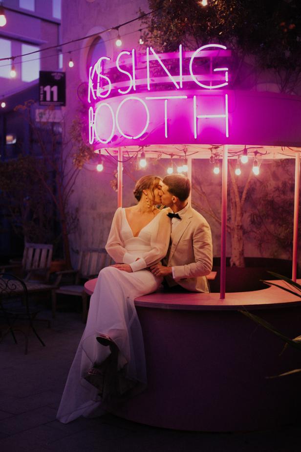 Ein Brautpaar küsst sich vor einem rosa „Kissing Booth“ mit Neonbeleuchtung.
