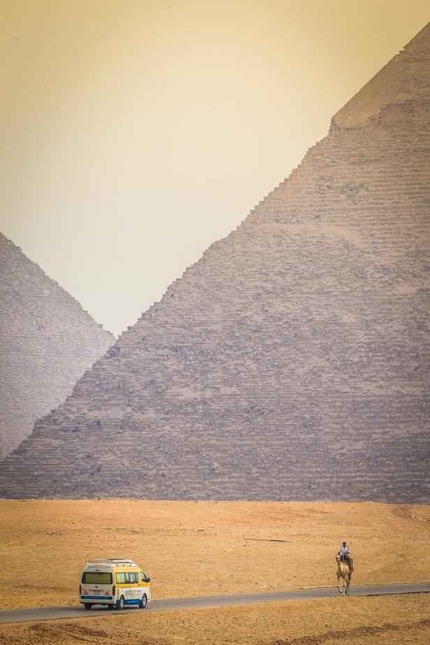 Ein Mann reitet auf einem Kamel auf einer Straße vor den Pyramiden von Gizeh, neben einem Touristenbus.