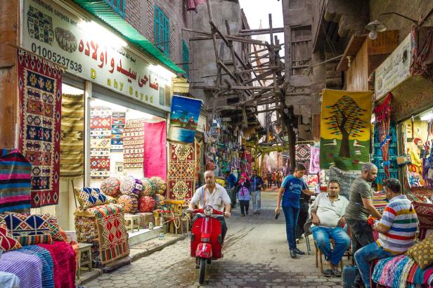 Ein Mann fährt auf einem roten Motorroller durch eine belebte Gasse mit Teppichgeschäften in Ägypten.