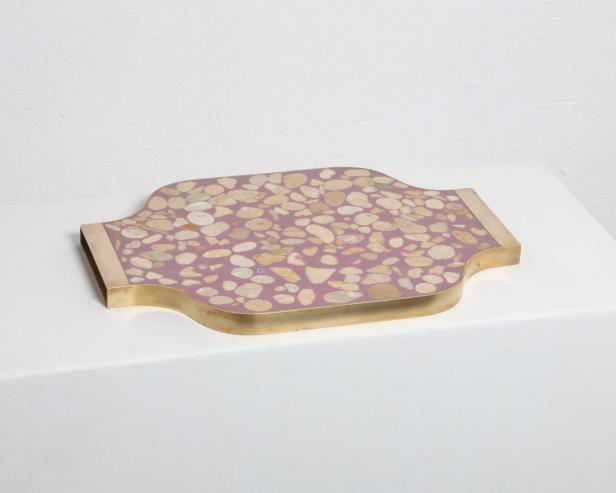 Ein dekoratives Tablett mit Terrazzo-Muster und goldenen Griffen liegt auf einer weißen Oberfläche.