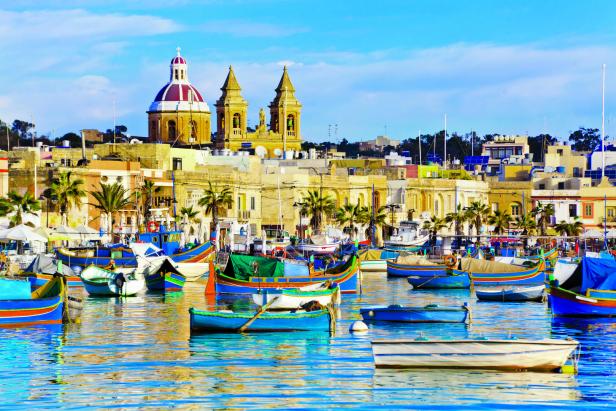 Bunte Fischerboote liegen im Hafen von Marsaxlokk, Malta, vor der Stadtkulisse.