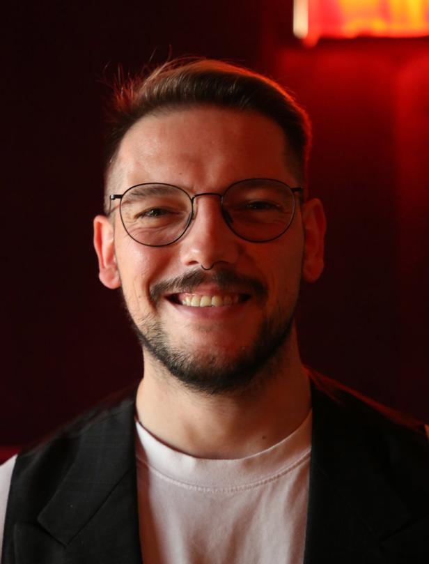 Ein lächelnder Mann mit Brille, Bart und Weste vor einem roten Hintergrund.