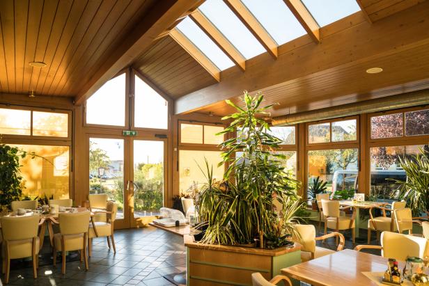 Ein lichtdurchfluteter Wintergarten mit Holzbalken und vielen Pflanzen, der als Restaurant genutzt wird.