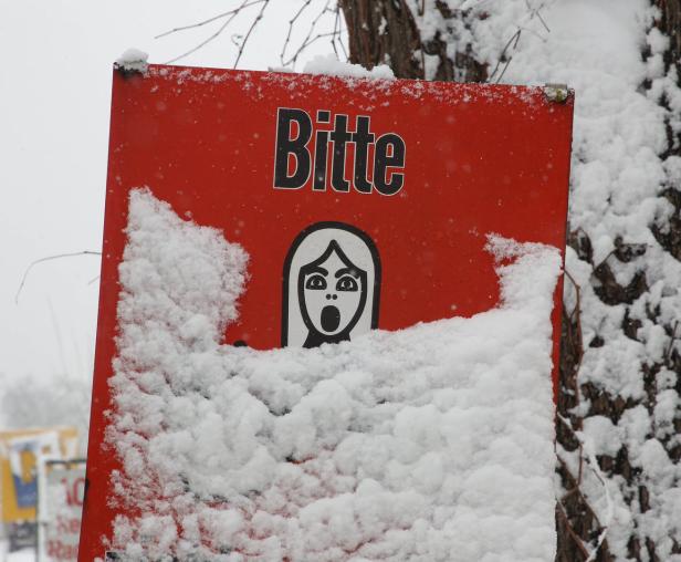 Ein rotes Schild mit der Aufschrift „Bitte“ und einem Piktogramm ist mit Schnee bedeckt.