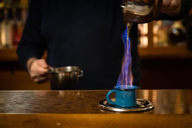 Ein Barkeeper gießt eine brennende Flüssigkeit in eine blaue Tasse.
