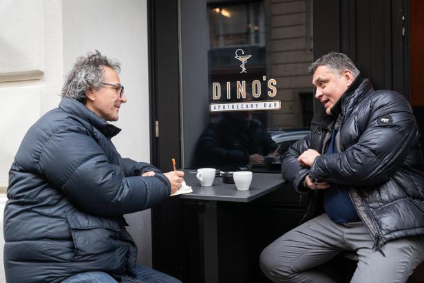 Zwei Männer sitzen an einem Tisch vor „Dino's Apothecary Bar“ und unterhalten sich.