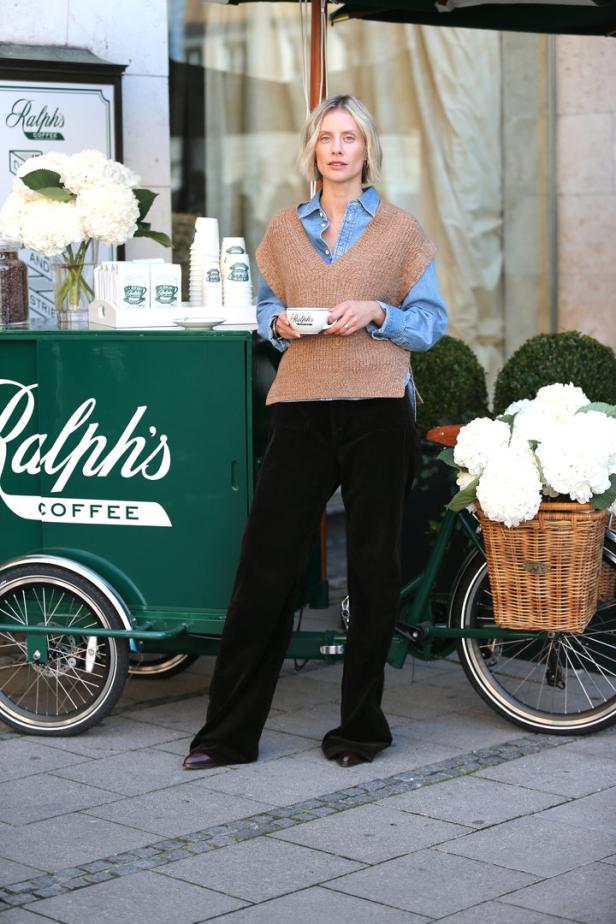 Eine Frau steht vor einem „Ralph's Coffee“-Stand und hält eine Tasse Kaffee.