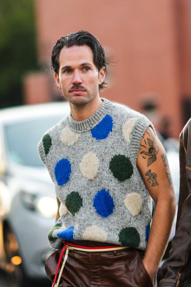 Ein Mann mit Schnurrbart trägt eine ärmellose Strickweste mit bunten Punkten und braune Lederhose.