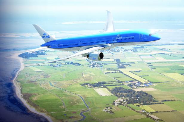 Ein blaues KLM-Flugzeug überfliegt grüne Felder und eine Küstenlinie.