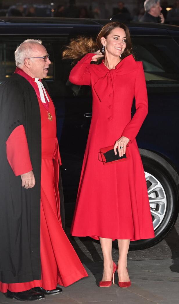 Kate Middleton in einem roten Kleid mit passenden Schuhen und Handtasche neben einem Geistlichen.