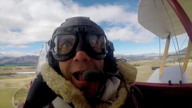 Ein Mann mit Fliegerbrille und -mütze in einem offenen Doppeldecker über einer Landschaft.
