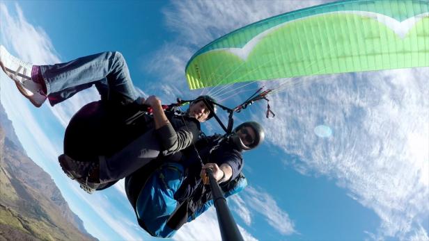 Zwei Männer machen ein Selfie beim Tandem-Gleitschirmfliegen vor blauem Himmel.
