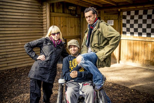 Drei Personen posieren vor einem Holzgebäude, eine davon im Rollstuhl.