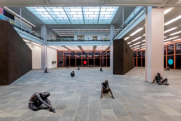 Eine Kunstinstallation mit sitzenden, dunklen Figuren in einem großen Raum mit Oberlicht.