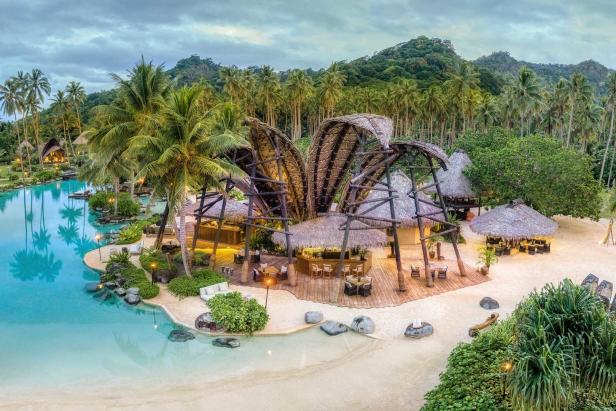 Ein luxuriöses Resort mit strohgedeckten Hütten am türkisfarbenen Wasser, umgeben von Palmen.