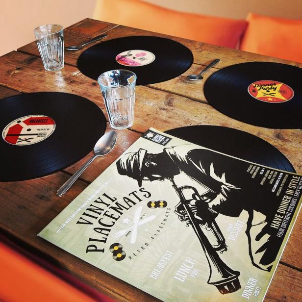 Ein Esstisch ist mit Schallplatten-Platzsets, Gläsern und Besteck gedeckt.
