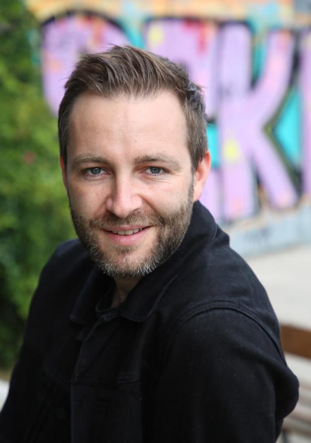 Ein lächelnder Mann mit Bart und dunkler Jacke vor einer Graffiti-Wand.