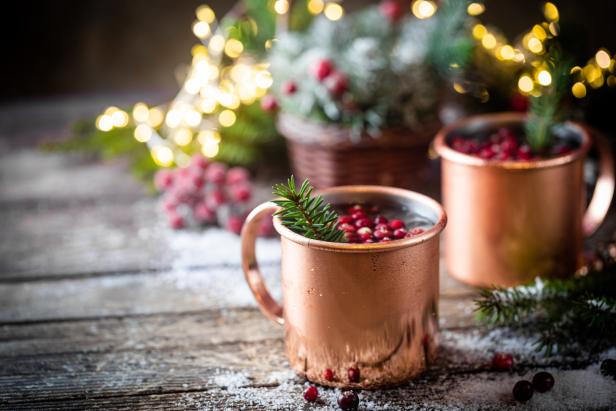 Zwei Kupferbecher mit Getränken und Preiselbeeren auf einem Holztisch mit weihnachtlicher Dekoration.