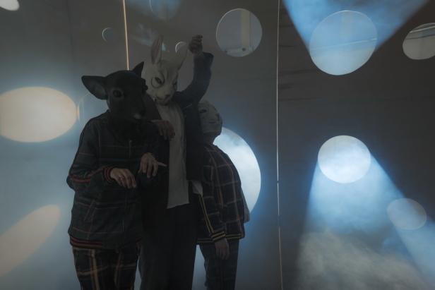Drei Personen mit Tiermasken stehen vor einer Wand mit kreisförmigen Öffnungen.