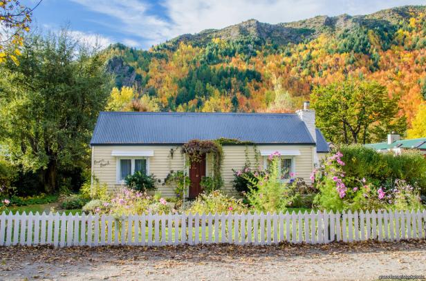 Ein gelbes Haus mit blauem Dach und einem Garten mit Blumen vor einem bewaldeten Hügel.
