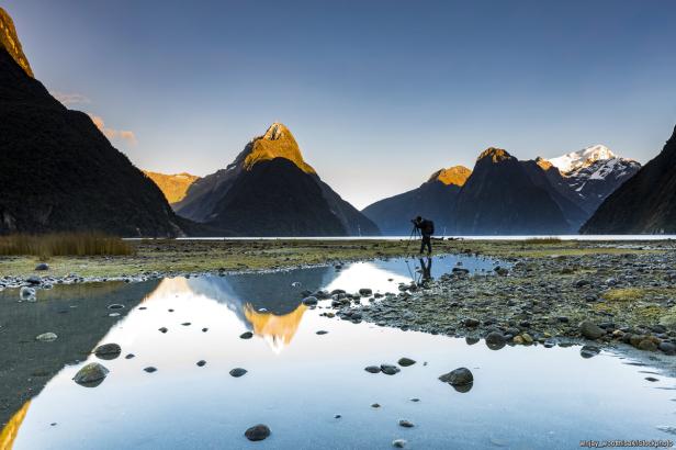 Ein Fotograf steht vor einer Bergkulisse und spiegelt sich im Wasser.