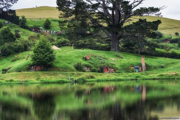 Blick auf Hobbiton mit kleinen Häusern in grünen Hügeln, die sich im See spiegeln.