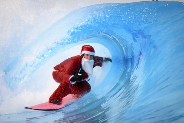 Ein Weihnachtsmann surft auf einem rosafarbenen Surfbrett durch eine blaue Welle.