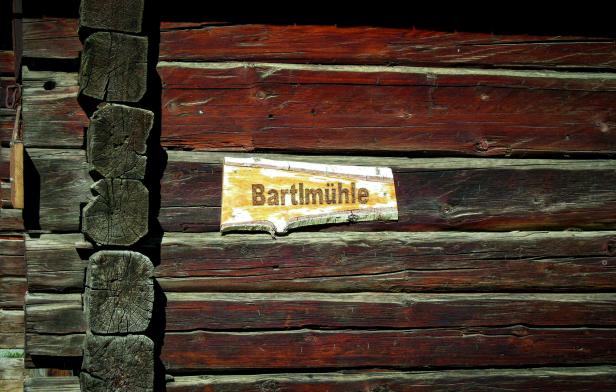 Ein Schild mit der Aufschrift „Bartlmühle“ an einer Holzwand.
