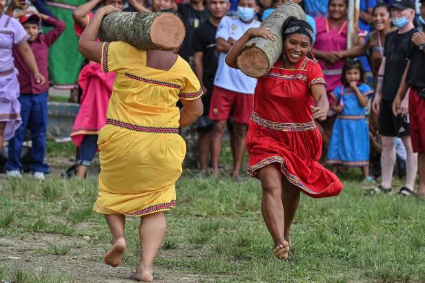 Zwei barfüßige Frauen in traditioneller Kleidung tragen Baumstämme auf ihren Schultern.
