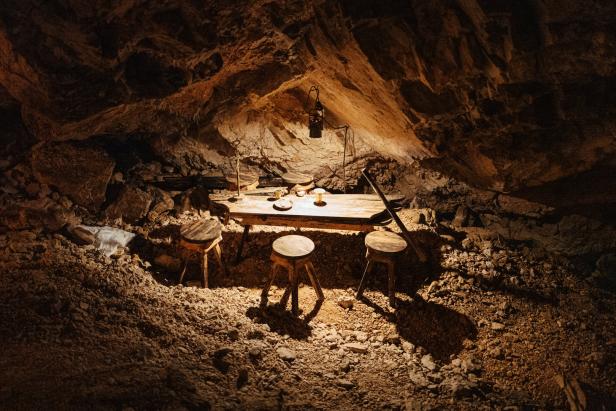 Ein Tisch und drei Hocker in einer dunklen Höhle, beleuchtet von einer Lampe.