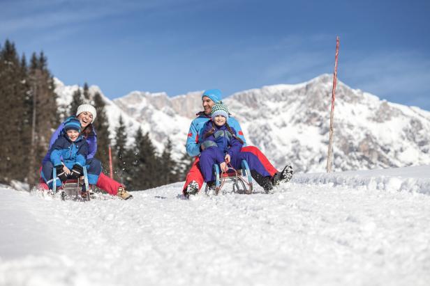 Eine Familie fährt auf Schlitten einen schneebedeckten Hügel hinunter, im Hintergrund schneebedeckte Berge.
