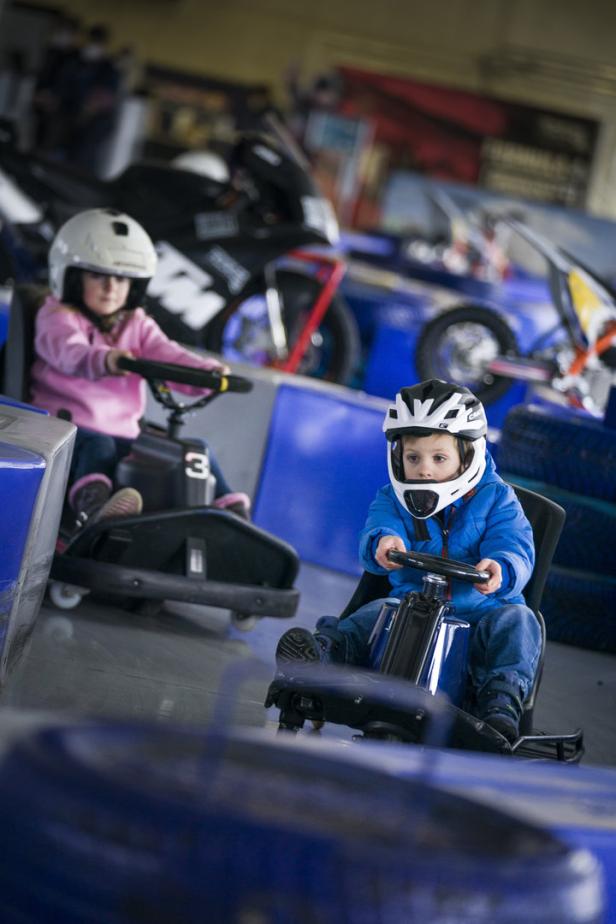Zwei Kinder mit Helmen fahren in kleinen Elektroautos auf einer Rennstrecke.