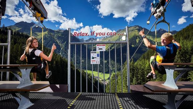 Eine Frau und ein Mann geben auf einer Zipline vor einer Bergkulisse ein positives Zeichen.