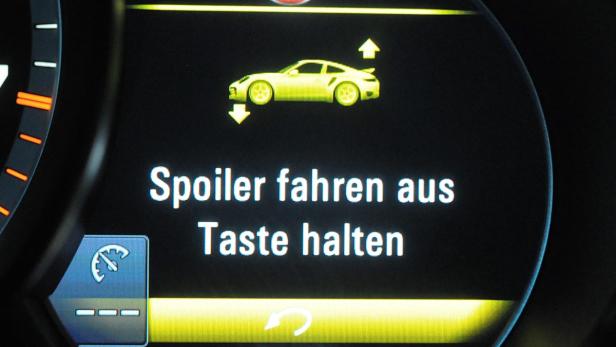 Das Display eines Autos zeigt die Meldung „Spoiler fahren aus. Taste halten.“.